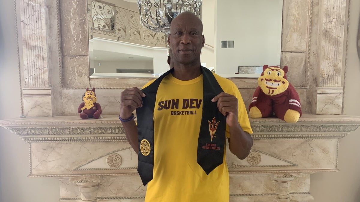 Man wearing a yellow "SUN DEVIL BASKETBALL" T-shirt holding a black graduation stole, standing in front of a marble mantel with plush mascots.