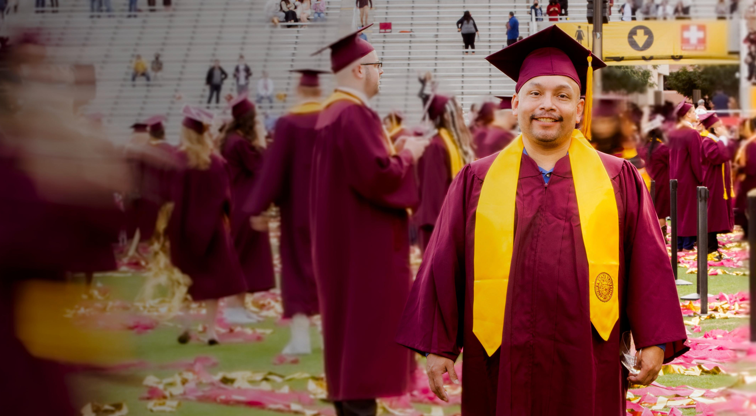 A man in a maroon cap and gown with a gold sash at a graduation ceremony, surrounded by other graduates and confetti.