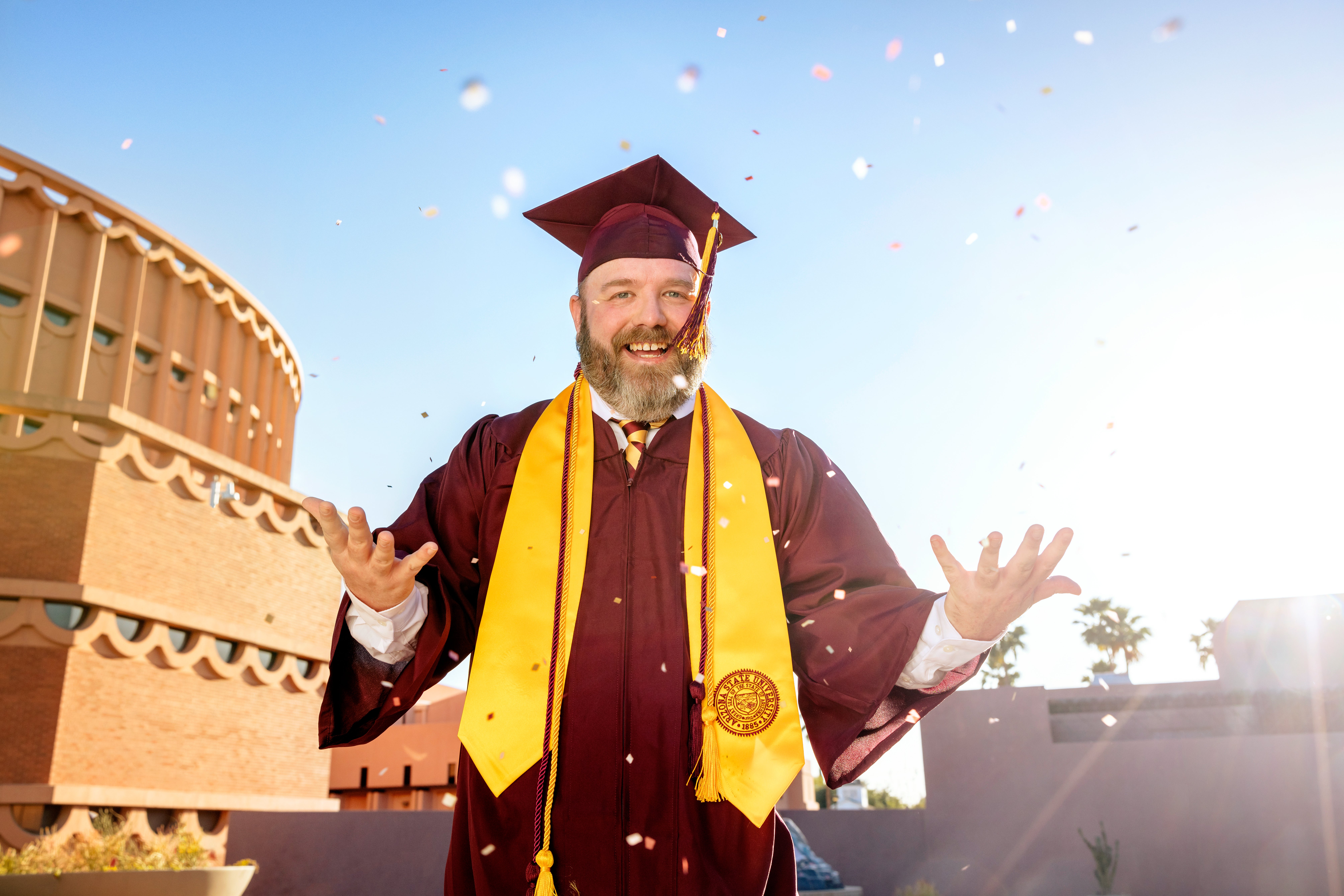 A joyful graduate wearing maroon and gold Arizona State University regalia throws confetti in the air while smiling. He stands in front of a campus building under a clear blue sky, with palm trees and sunlight in the background.