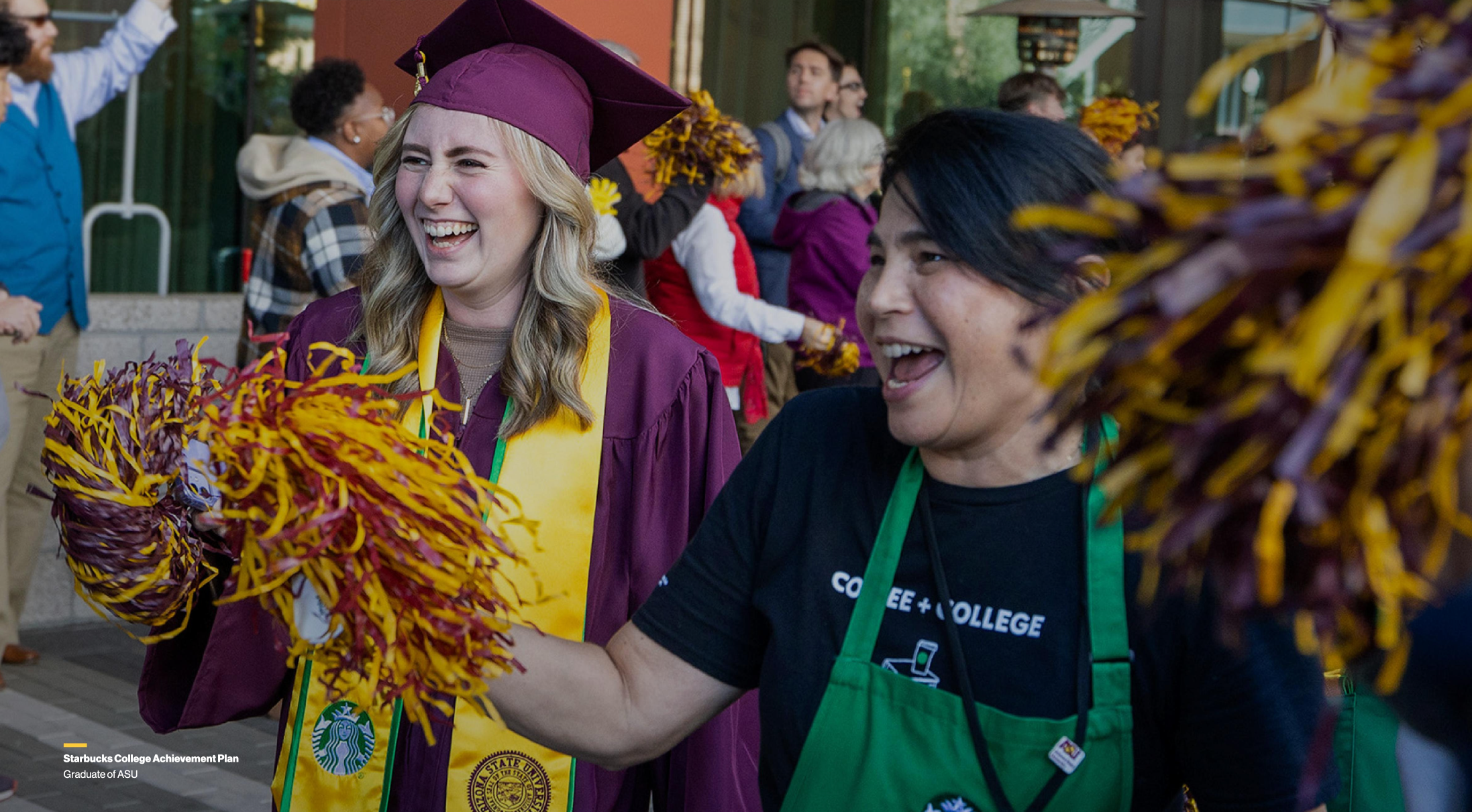 A graduate in a maroon cap and gown celebrating with pom-poms alongside another person in a green apron.